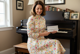Woman in a floral dress holding a lyssy may sheet music wallet in front of a piano.