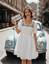 Woman in a white dress with floral embroidery holding a lyssymay street map wallet, standing in front of a vintage car on a street.